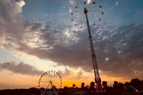 Carl A ferris wheel and a towering fairground ride dominate this image. Behind them lies a cloudy sky with a rising sun beginning to cast it's golden light across the view.