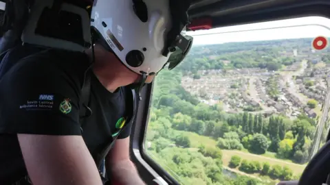 A white middle-aged man in a black t-shirt, wearing a white crash helmet, sitting in a passenger seat on a helicopter, looking out of a window down to park and rooftops of houses