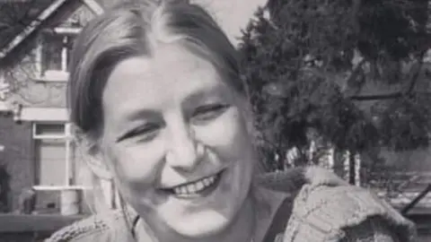 Caroline Sturgess A black and white photo of Dawn Sturgess who is smiling as she sits outside by some trees and buildings