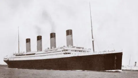 A black and white photo of RMS Titanic, a large ocean liner with four funnels, a black hull and a white upper deck. There are masts at the front and rear.