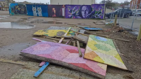 a muddy car park with painted boards strewn on the ground, artworks on upright boards and large puddles in the background with metal fencing and cars to the right 