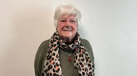 A woman with grey hair and wearing a green jumper, a spotty scarf and a large amber necklace, smiling at the camera against a white background.