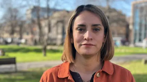 Samira Dehghanazad is standing in a green space bordered by buildings, and looking at the camera with a neutral expression on her face. She has short light brown hair and is wearing an orange jacket.