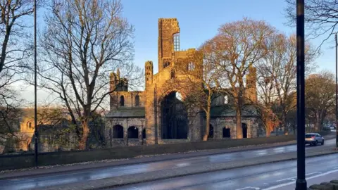 A ruined abbey on a cold winter's day. It is surrounded by trees and viewed from the opposite side of a main road.