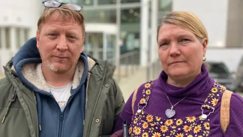 The image shows two people standing outdoors in what looks like a car park or a paved area with several parked cars behind them. One person is wearing a dark green padded coat with a hood and glasses, while the other is wearing a purple top with a patterned dress or pinafore and carrying a bag over one shoulder.