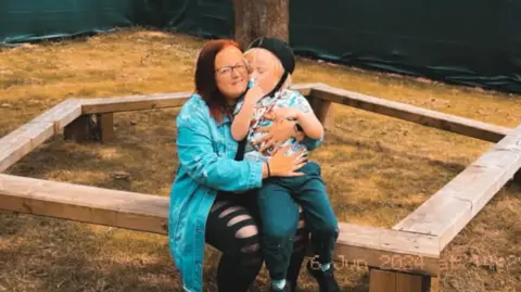 Family A woman with red hair and glasses sits on a wooden bench in a garden, holding her young son on her lap. The boy is wearing a baseball cap and a patterned shirt, leaning into his mother. A large tree stands behind them, with a green fence and houses visible in the background.