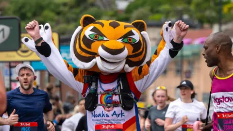 A J Bell Great Bristol Run A person running as they wear a Noah's Ark children's hospital charity top and a cartoon tiger head. There are other people each side and behind them, in sports gear, running the Great Bristol Run.