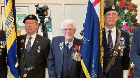 GUY CAMPBELL/BBC Mr Grant, displaying his medals on his blue jacket, stands between two standard bearers of the Royal British Legion holding flags by their side