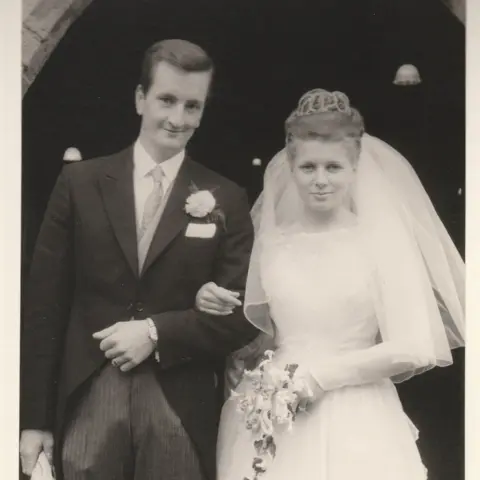 Family A black and white photo of a groom and bride on their wedding day. He wears a suit with a flower in his lapel while the bride wears a veil, white dress, and holds a bouquet. Their arms are linked and they smile at the camera.