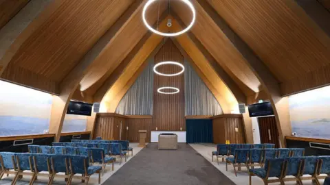 Interior of Halo Hall. A large modern hall with a vaulted wooden ceiling, large pictures on the walls and rows of blue velvet chairs.