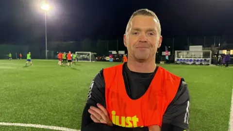 A man in a football kit and bright red bib stands on the edge of a flood lit astro turf pitch with his arms folded and smiling at the camera