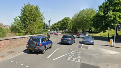 Google Cars driving on the four-way junction at Maumbury Cross in Dorchester. To the left is a brick wall with a narrow pavement.