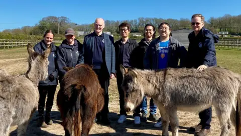 Isle of Wight Donkey Sanctuary/University of Southampton A group of seven people looking at the camera, including staff from the donkey sanctuary and scientists from the University of Southampton. There are 3 donkeys in front of them and fields and fences behind them.