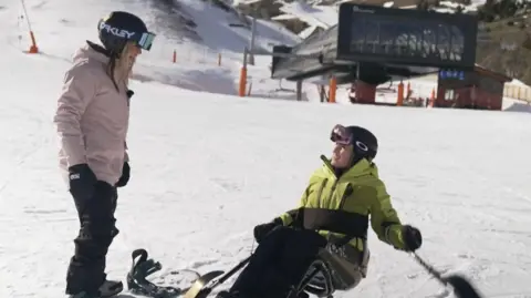 Ellie sitting on her sit-ski on the white snowy mountains in Andorra. She wears a green-yellow ski coat, a black helmet and sun visor. Across her middle is a black strap and behind her is a ski lift building. She is looking up and talking to Jenny Jones, Ski Sunday presenter, who wears black ski trousers, a pale pink jacket and black helmet