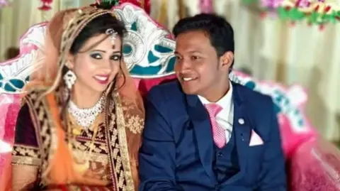 BBC The image is of an Indian couple, with the groom wearing a suit and the bride dressed in traditional Indian wedding attire with jewellery.