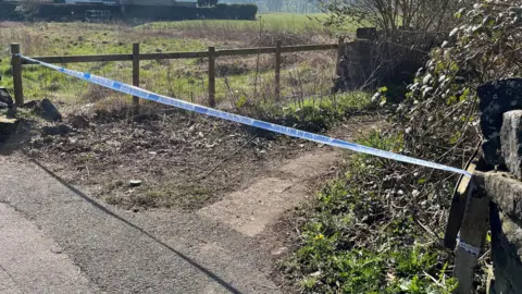 Police tape on the entrance to a small lane leading into a wooded area.