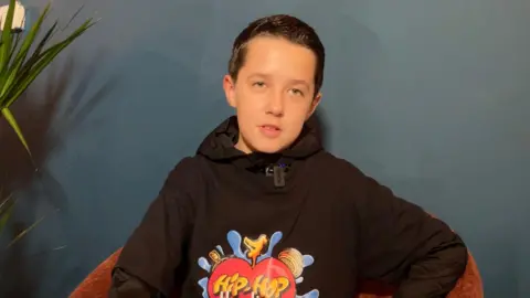 A picture of Kade sitting in a dark red chair in front of a dark blue wall, next to a plant on the left. He wears a black hoodie with a black shirt with a colourful Hip-Hop Healing graphic on top of it.