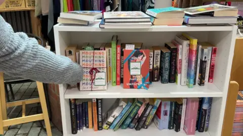 A book shelf. There two shelved are filled with books. There is an arm, grey sleeve, putting a book on the top shelf, with a free book bookmark sticking out of the book and tied with red strong and with a book fairy sticker on. To the right of it there is another book with a book fairy sticker and blue ribbon and free book sign sticking out. On the of the book shelf are books lying flat. 