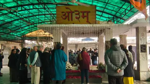 BBC Hindi People gather around a platform which is covered with flowers in a crematorium in Delhi. This is a scene from Mark Tully's cremation. 