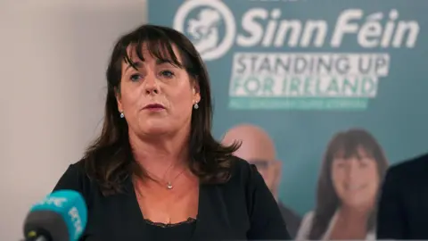 Michelle Gildernew speaking at the launch of the party's manifesto for the European election campaign in May 2024.  She has shoulder-length dark hair with a fringe and is wearing a black top under a black jacket along with a necklace and earrings.   She is speaking into a green microphone and there is a green Sinn Féin poster with her face on it behind her. 