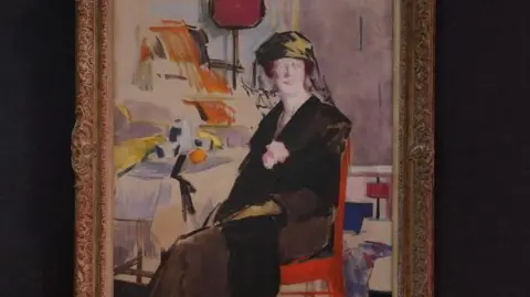 An oil painting of a woman wearing black sitting on a chair. There are other pieces of furniture in the background. The painting is framed and hung on a black wall.