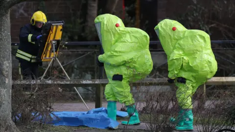 PA Media Two people dressed in large, neon, yellow hazmat suits. Walking on a sidewalk in front of a tree on a gray day. A firefighter in the background with equipment.