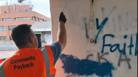 Swindon Borough Council A man in an orange high vis jacket which says 'community payback' paints over some graffiti on a white wall