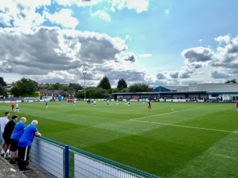BBC Weather Watchers/Beefy Several men in Halesowen lean on a railing next to a green football pitch with white lines marking out the pitch. Several players can be seen in the distance.