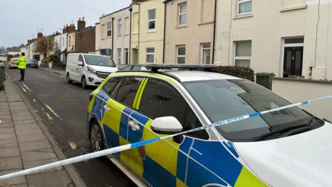 BBC Blue and white police tape cordons off a residential street of terraced houses. A police car is in the foreground and an officer can be seen down the road. 