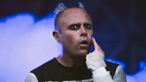 Keith Flint is on stage while wearing a black top with white sleeves. He has dark eyeliner on and his head is shaved, except for two parts which stand up like devil horns. Blue smoke billows behind him.