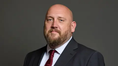 House of Commons A bald man with a beard wearing a black suit and red tie.