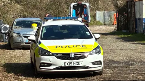A high visibility police car is parked in a area by a train station. Behind it is a silvery-grey car, with a yellow item on its bonnet, and in the background is a van with its back doors wide open.