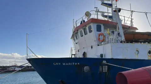 Scilly Steamship Group The Gry Maritha, with a blue base and white cabin, is seen approaching from the right with the sea beyond. 