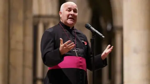 Stephen Cottrell, Archbishop of York, giving a sermon dressed in black with a red belt, wearing a poppy and a cross necklace. He is balding with white hair.