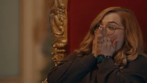 Courtesy of Netlix Sarah Taylor, a woman in her thirties, sits on a throne-like chair with her hands covering her face in disbelief. 