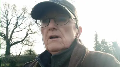 Lesley Martin has short hair, a black cap, thin black oval shaped spectacles and is wearing two fleece jackets. The image is in hazy sunshine.