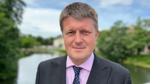 Man wearing suit, purple shirt and tie, looking at the camera with a blurred lake and trees behind him