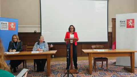 Michael Keohan/BBC Linden Kemkaran wearing a red jacket, stands at a lectern during a presentation at County Hall in Maidstone.