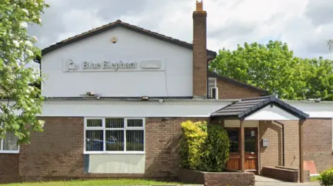 A brick building with a tall chimney and white wooden cladding on the first floor. There is a silver sign which reads Blue Elephant on the side