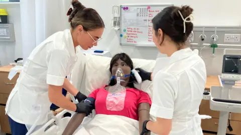 Two female students in white tops either side of a mannequin on a hospital bed. The nurses both have long dark hair tied back and the mannequin is wearing a pink t-shirt and a mask on its face.