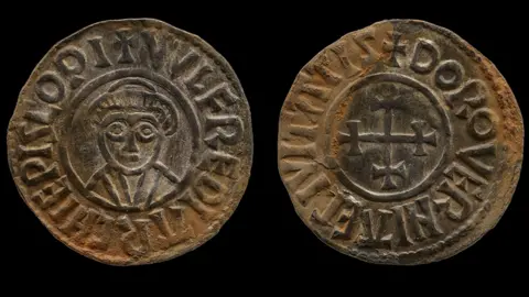 A silver ingot coin which has shows a man on one side and a cross on the other