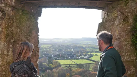 National Trust/ Richard Gregory A woman on the left and a man on the right both peering out of a lookout from the old castle walls. Fields, housing and trees can be seen in the distance as they look out on the scene.