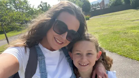 A selfie image of a woman stood in a park in the sunshine with her arm around a young girl. They are both wearing white t shirts and blue denim dungarees. They both have wavy brown hair and the woman is also wearing sunglasses and a grey backpack over one shoulder. behind them is grass and trees. They are both smiling and looking at the camera.