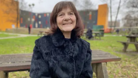 Hazel, 65, has a brown bobbed haircut and is wearing a black fluffy coat. She is sitting on a picnic bench in the grounds of Cardiff's University Hospital of Wales.  
