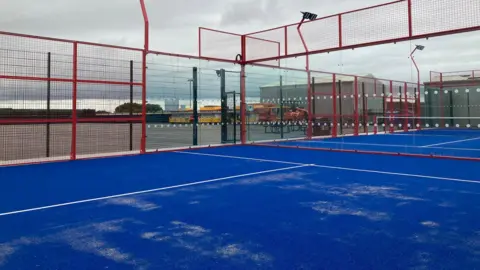 BBC A blue floored padel court, surrounded by red fences. 