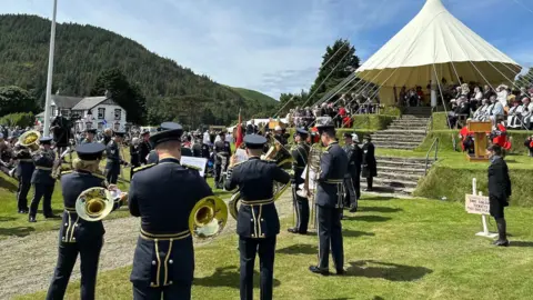 BBC A military band playing in front of the wedding cake hill with officials sat on it.