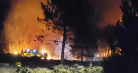 A large fire illuminates the night sky, as firefighters battle around it to get it under control.