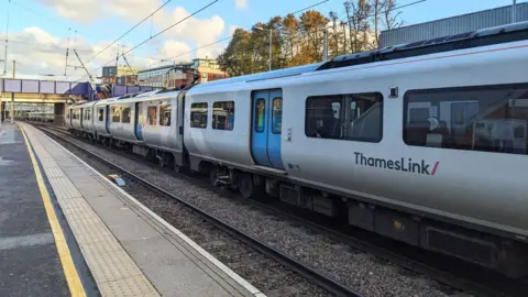 A Thameslink train stopping at West Hampstead Thameslink Station with a footbridge at the back next to an empty platform under a blue sky during sunset