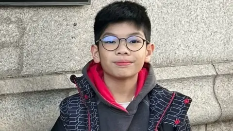 SCOTLAND POLICE Thomas Wong looks directly at the camera. He has large glasses on his face and dark hair. He wears a gray hoodie with a red interior lining and a black jacket with a red zipper and button and a white box design. He stands in front of a light stone wall.