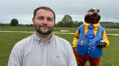 Max Biddlestone is pictured in a blue and white striped shirt. He is standing in front of a mole statue in a field. The statue is wearing a bright yellow shirt, a blue tie, blue waistcoat and red trousers.